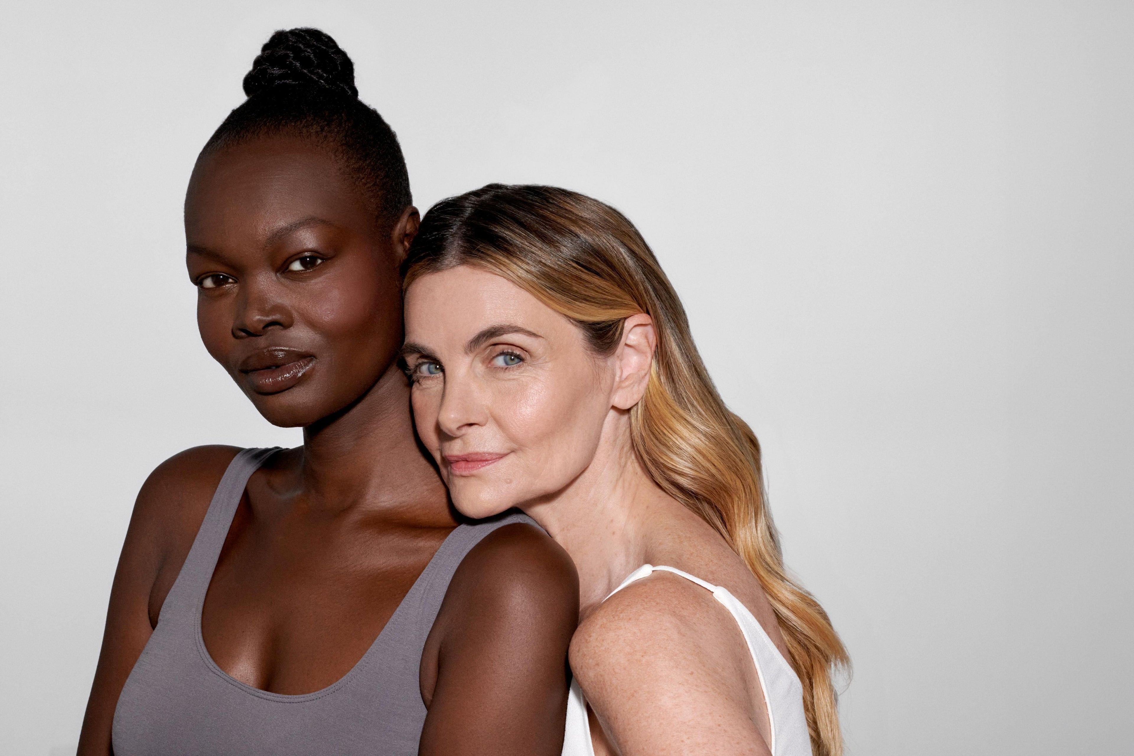 Two women with healthy, glowing skin standing close together against a neutral background, showcasing natural radiance across different skin tones.