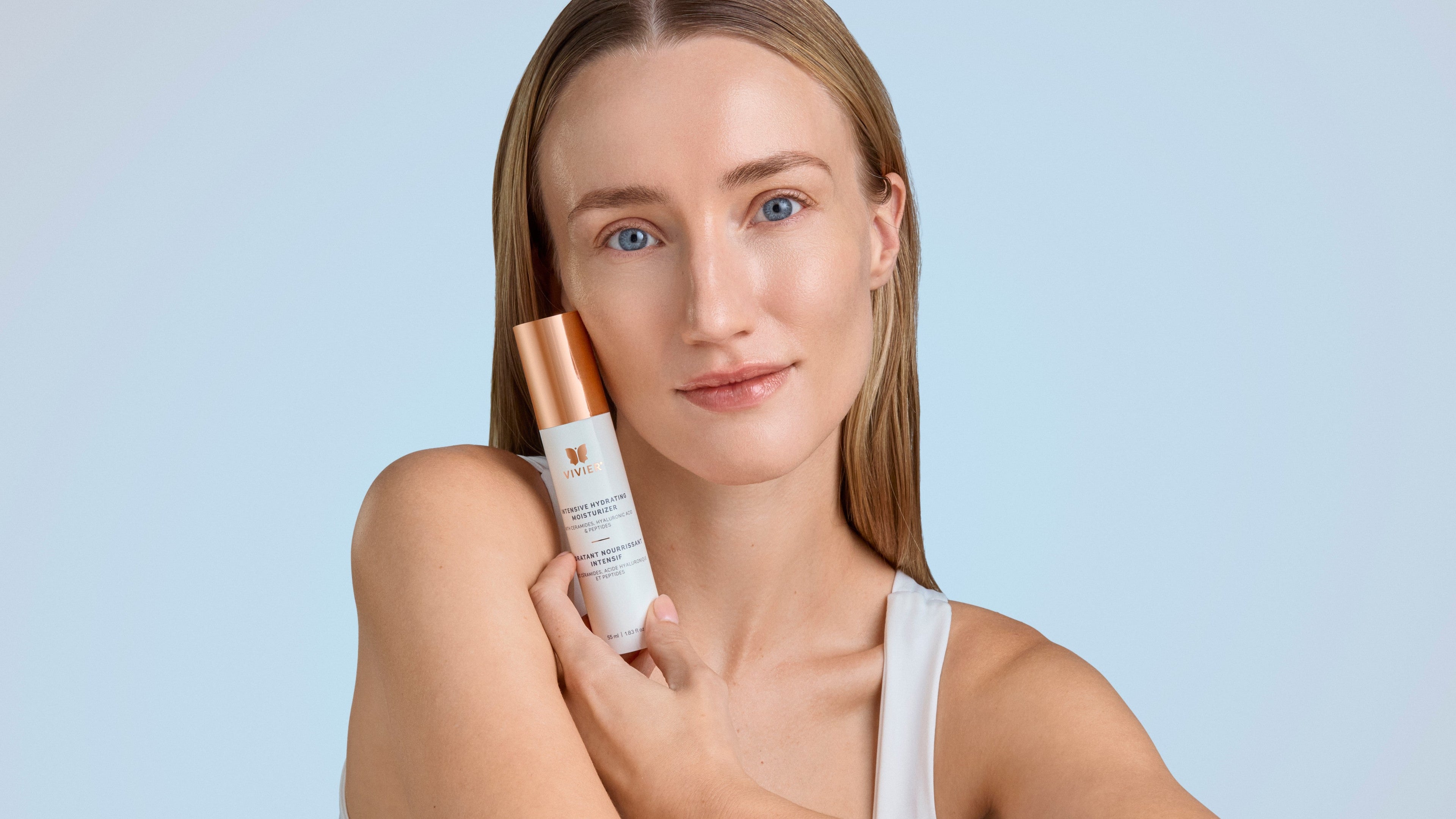 A woman with natural, radiant skin holds Vivier’s Intensive Hydrating Moisturizer against her cheek, set against a soft blue background with clean, minimal styling.