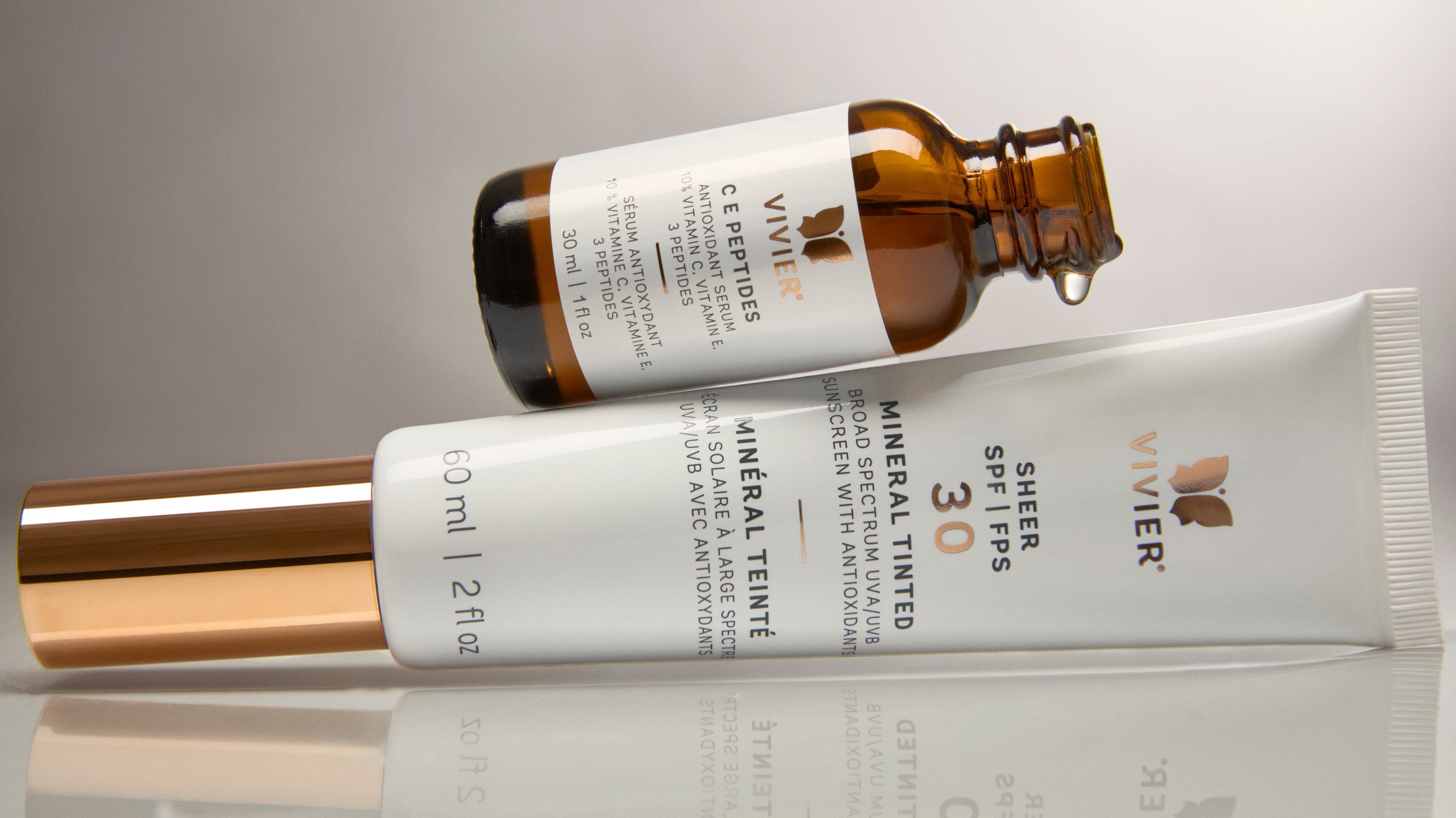 Flat lay of Vivier C E Peptides serum bottle and Sheer SPF 30 Mineral Tinted sunscreen on a reflective surface with soft neutral lighting.