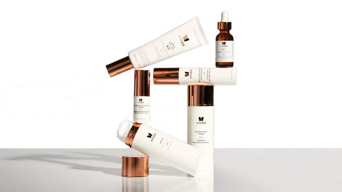 Alt text: Vivier skincare products arranged in a balanced display against a clean white background, featuring sunscreen, vitamin C serum, toner, eye cream, and cleansing products with signature rose gold packaging.