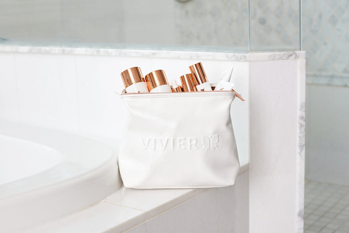 White Vivier skincare bag filled with products, placed on a bathroom counter beside a bathtub